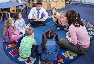 stock-photo-group-of-nursery-children-sitting-on-the-floor-in-their-classroom-the-male-teacher-is-reading-from-447240136